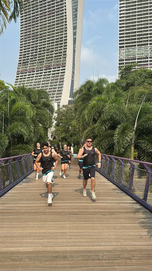 A group of friends jogs across a wooden bridge in Singapore, with the iconic Marina Bay Sands towers rising in the background. The image captures the lively morning run referenced in the caption, set against the city's lush greenery and modern skyline.