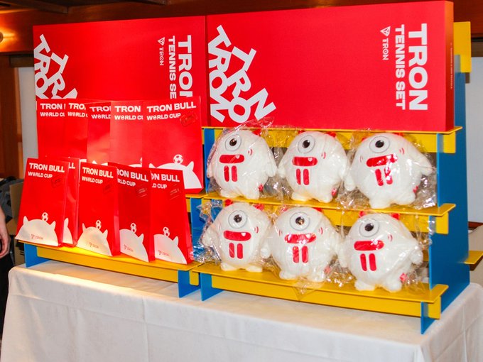 A display table features red "TRON TENNIS SET" boxes alongside "TRON BULL WORLD CUP" packages, with six white plush bull mascots wrapped in plastic arranged on yellow and blue shelves—souvenirs from the TRON Global Friends JAPAN 3rd offline event.