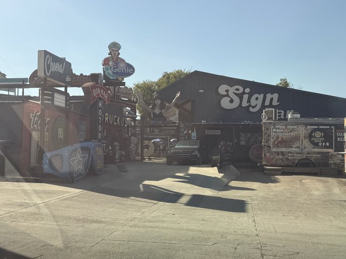 An outdoor vintage sign lot featuring a large 'Sign' storefront, a colorful 'Genie' cutout, a 'Cheers' sign, and various other old business signs and a blue car hood with a star.