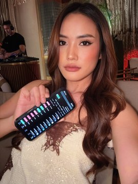 A young woman with long brown hair and pink eyeshadow holding up a smartphone displaying a financial/stock trading chart with green and red candlesticks.