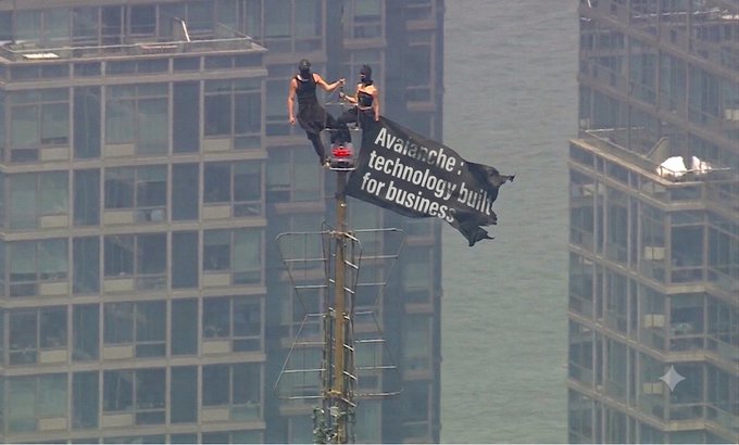 Two activists climb a communications tower holding a banner reading 'Avalanche: technology built for business' against an urban skyline backdrop.