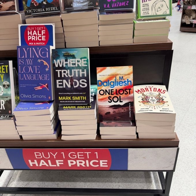 A bookstore display table promoting a 'BUY 1 GET 1 HALF PRICE' sale, featuring prominently displayed books including 'Lying Is My Love Language' by Olivia Simons, 'Where Truth Ends' by Mark Smith, 'One Lost Soul' by J.M. Dalglish, and 'Morton's.'