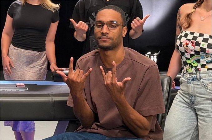 A man wearing glasses and a brown t-shirt sits at a table flashing the "rock on" hand gesture with both hands, while two women standing on either side also make similar hand signs. The image humorously embodies the post's "habibi bagwork" celebration vibe, capturing an enthusiastic, almost ritualistic pose reminiscent of crypto traders reveling in a Bitcoin "god candle" price surge.