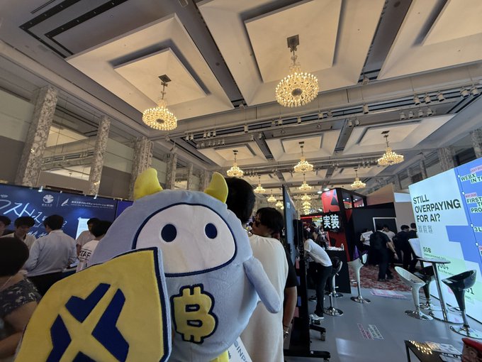 A photo taken at a conference or event showing a person in a white and yellow mascot costume with a Bitcoin logo on it, with booths including one that says 'STILL OVERPAYING FOR AI?' visible in the background.