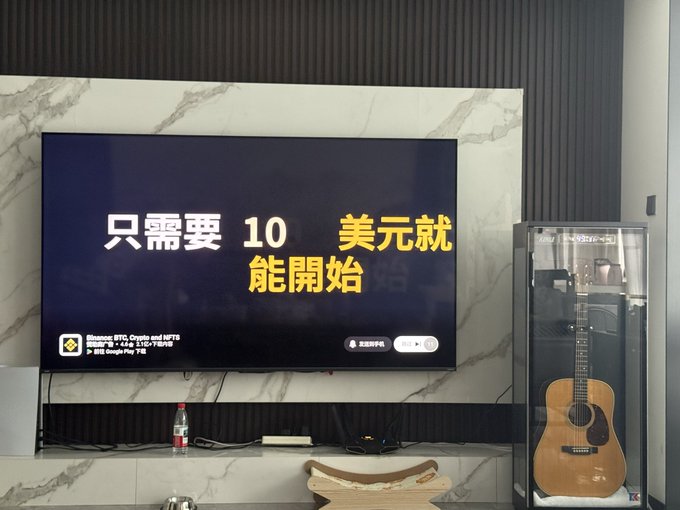 A wall-mounted TV displays Chinese text '只需要10美元就能開始' (Start with just $10) alongside an advertisement for 'Bitcoin (BTC), Crypto, and MPT8' on BTCC, with a Martin acoustic guitar visible in a glass display case to the right.