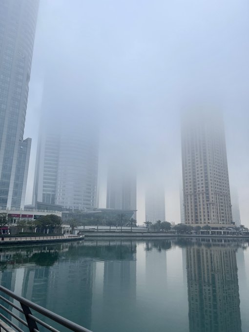 A foggy cityscape with tall skyscrapers partially obscured by mist, their reflections visible in the calm water below.