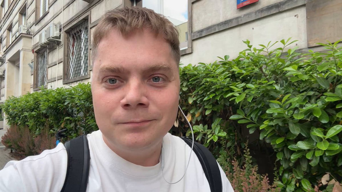 The image features a man wearing a white shirt with headphones around his neck, standing next to some bushes outside of an old building. The man appears to be looking directly at the camera, possibly posing for a photo or recording a video. The surrounding area includes a sidewalk and a street, suggesting that this scene takes place in an urban setting.
