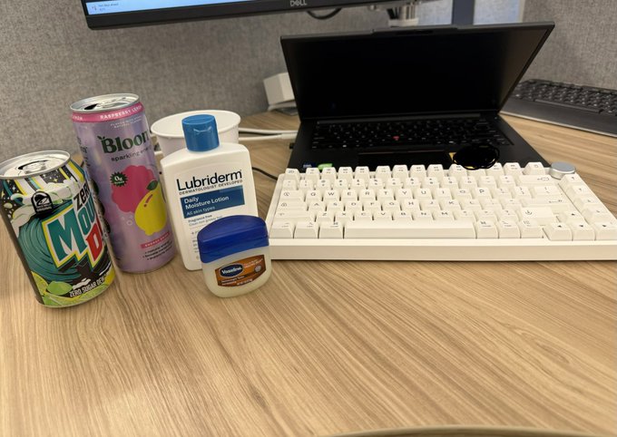 A desk setup showing Mountain Dew, Bloom energy drink, Lubriderm lotion, and Vaseline next to a laptop and white keyboard.