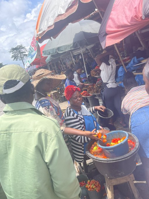 A bustling outdoor market scene where a vendor in a striped shirt serves tomatoes from a large metal bowl under colorful umbrellas.