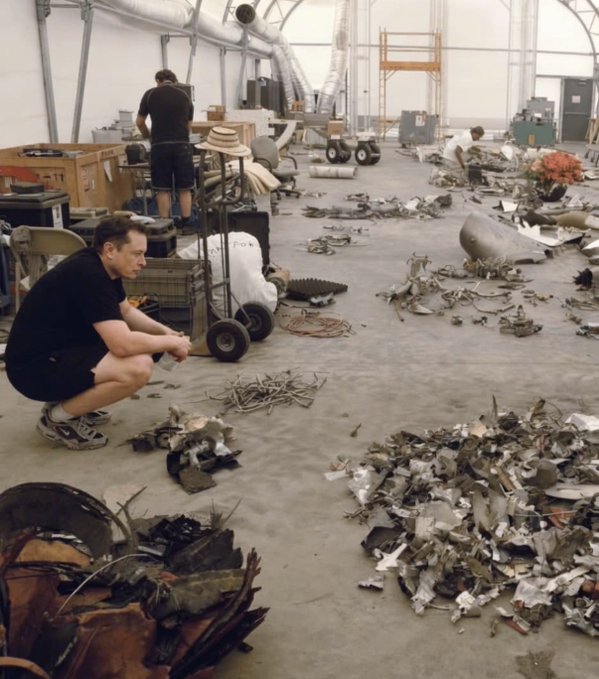 The image shows a man kneeling down next to a pile of scrap metal and other debris. He appears to be examining or sorting through the items. The surrounding area also contains various pieces of junk, including a chair and several boxes scattered around. A person is standing in the background, possibly observing the scene or working on their own task.