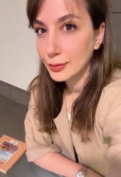 A selfie of a young woman with long brown hair wearing a beige top, posing indoors with what appears to be a book nearby.