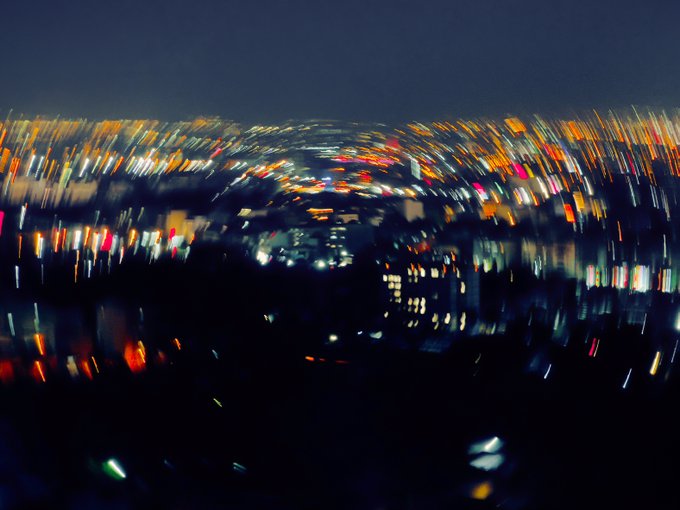 A heavily blurred nighttime cityscape photo featuring streaks of orange, yellow, white, and red lights from buildings and streets against a dark sky, creating an abstract motion effect.