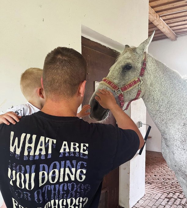 A man holding a small child greets a gray horse wearing a red halter, with the man's shirt reading "WHAT ARE YOU DOING" in bold white and blue lettering. The tender moment of bonding with the horse reflects the post's warm greeting of brotherhood and connection, treating the animal as a fellow "brother" in the family of life.