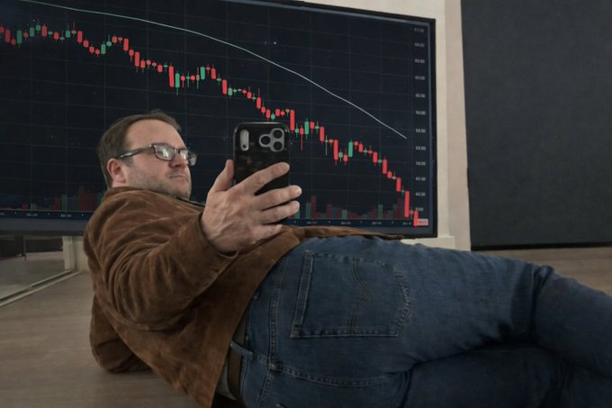 A man with glasses lies on the floor looking at his phone in front of a large monitor displaying a steeply declining stock chart, seemingly reflecting the bearish "$LIT" price action as he awaits news of its integration into the Robinhood Chain.