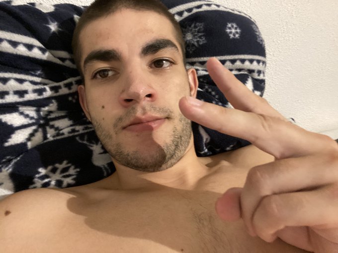 A shirtless young man with a short beard lying against a blue and white patterned pillow, making a peace sign with his fingers.