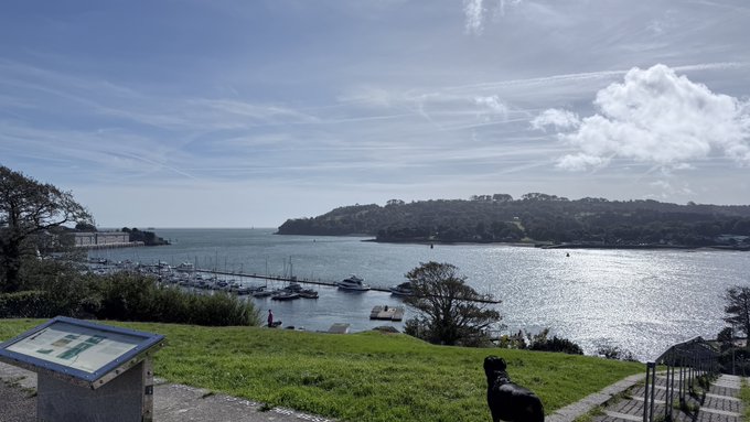 A scenic coastal view featuring a harbor with boats, a green hillside, and a body of water under a blue sky with scattered clouds.