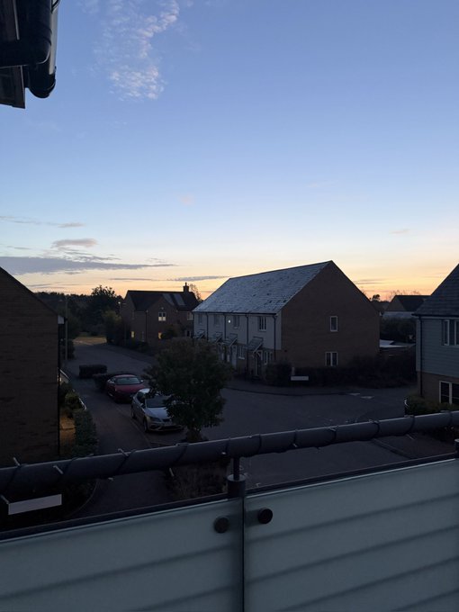 A serene twilight view from a balcony overlooking a suburban neighborhood with brick houses, parked cars, and a colorful sunset sky.