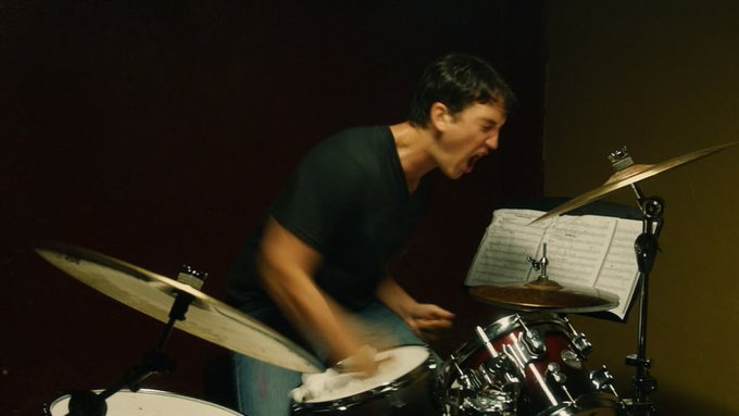 A man in a dark t-shirt plays a red drum kit energetically in a dimly lit room, with sheet music visible on a stand.