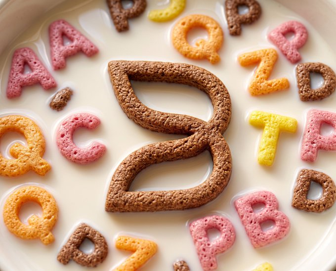 A close-up of a bowl of milk with colorful alphabet cereal floating on the surface, prominently featuring a large, textured brown letter 'B' in the center.