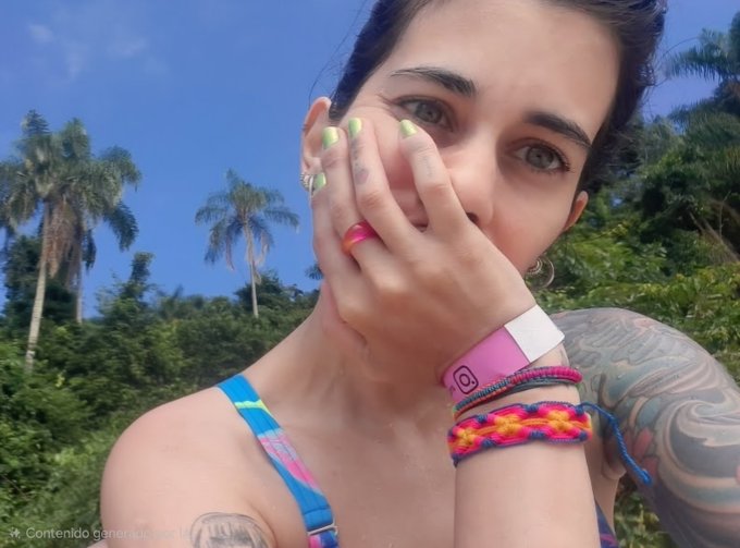 A close-up selfie of a young woman with light green nail polish covering half her face, wearing a colorful woven bracelet, an Instagram wristband, and rings, with a tropical background of palm trees and blue sky.