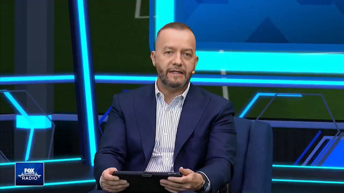 A bearded man in a blue blazer and striped shirt sits in a studio holding a tablet, with a "FOX SPORTS RADIO" logo visible in the bottom left corner against a blue-lit geometric background.