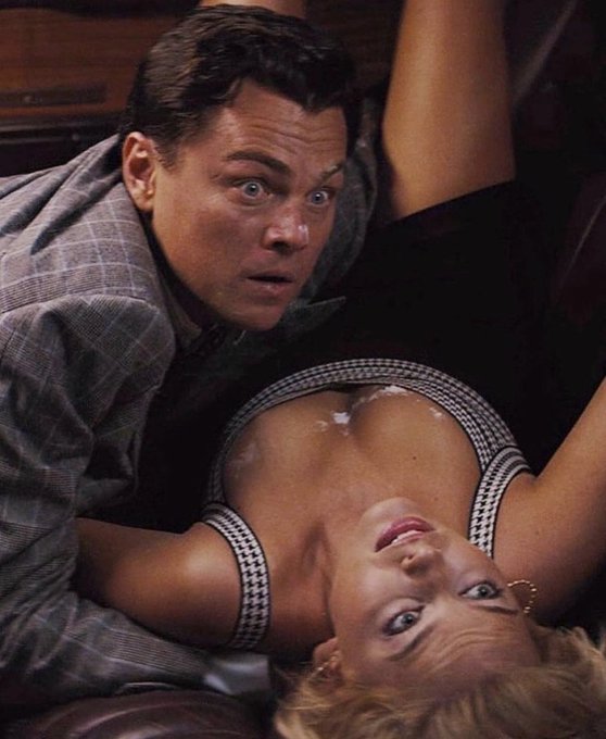 A scene from the movie 'The Wolf of Wall Street' showing Leonardo DiCaprio with a surprised expression looking down at Margot Robbie lying beside him.