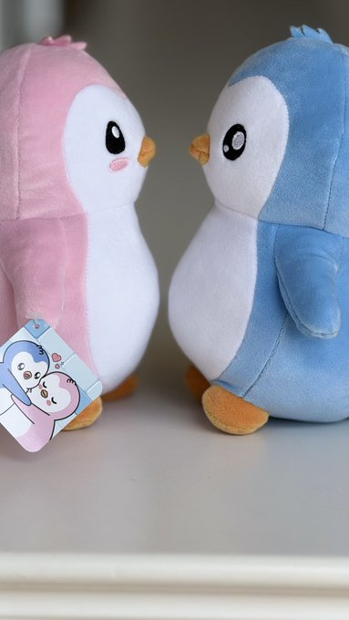Two plush penguin toys facing each other, one pink and one blue, with a heart-themed tag attached to the pink one.