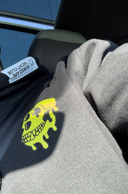 A person holds a bouquet of pink flowers while wearing a gray sweatshirt featuring a yellow melting skull graphic with X eyes, sitting inside a car.