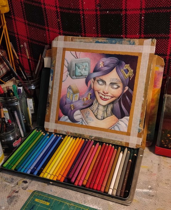 An open tin of colorful colored pencils arranged in rainbow order, with a detailed colored pencil drawing of a purple-haired character displayed in the lid.