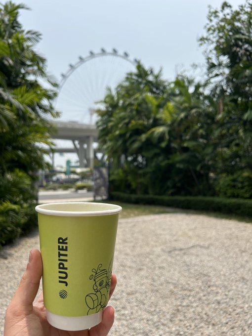 A hand holding a green paper cup labeled 'JUPITER' with tropical foliage visible and a large Ferris wheel in the background.