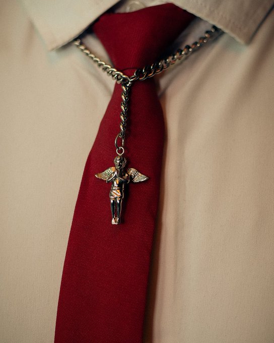 A close-up photograph of a deep red necktie worn with a beige dress shirt, accessorized with a silver chain necklace holding a small angel pendant resting on the tie.