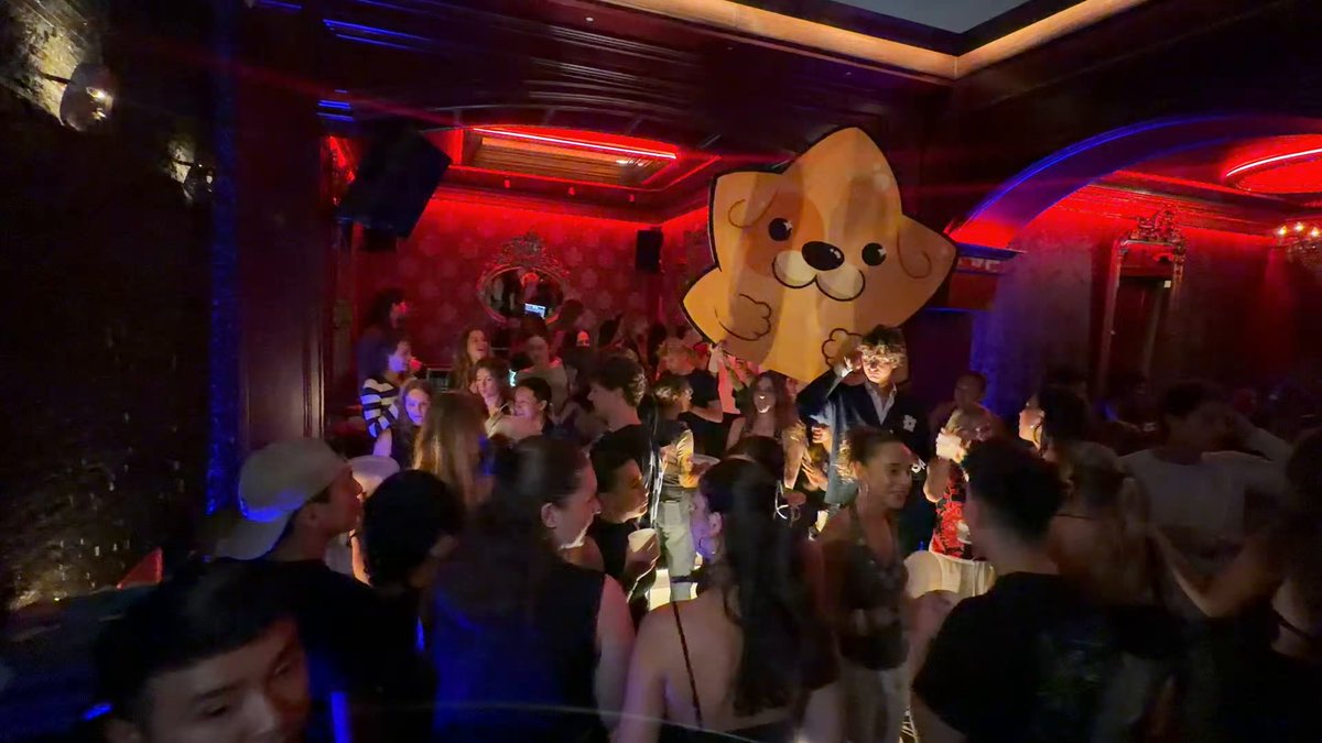 The image shows a large group of people gathered together, possibly at a party or event. The crowd appears to be enjoying themselves as they stand around each other. In the center of the scene, there's an oversized cartoon character resembling a dog that has been cut in half and is holding onto one end of its body with both hands. This unique decoration adds a fun and lively atmosphere to the gathering.