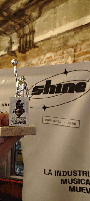 A photograph of a hand holding a small silver statue of a person with a raised fist, standing on a base that reads 'WEB3 STARTUPS LATINOAMÉRICA 2024,' held up in front of a white banner that says 'shine' and 'PRE-SEED · 2026' with 'LA INDUSTRIA MUSICAL' partially visible at the bottom.