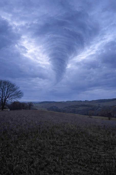 A dramatic funnel cloud descends from stormy gray clouds over a rural landscape with bare trees and a plowed field.
