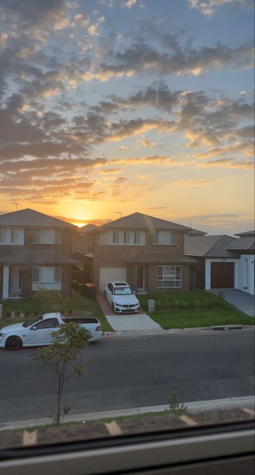 A suburban neighborhood at sunset with brick houses, a parked white BMW, and dramatic clouds lit up in orange and pink.