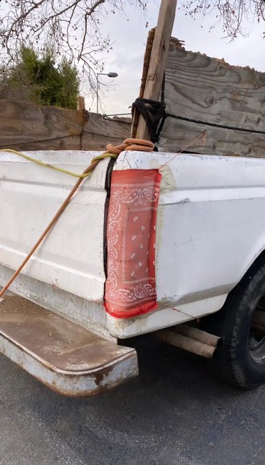 The back of a white pickup truck carrying wooden planks, secured with bungee cords and a red patterned bandana hanging from the side.