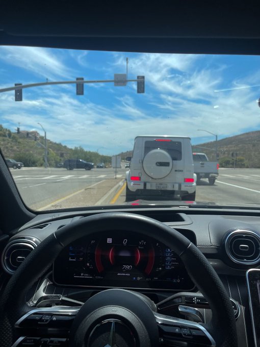 A driver's view from inside a Mercedes-AMG vehicle, following a white Mercedes-Benz G-Class SUV on a highway under a blue sky.