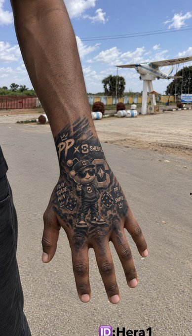 A photograph of a hand with a detailed black and grey tattoo featuring a bear character, dice, and text like 'PP', 'PLAY TRADE WIN', and 'SHUFL', watermarked with 'Hera1'.