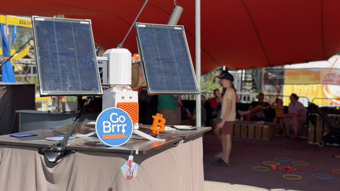 A solar-powered Bitcoin mining setup featuring two photovoltaic panels angled toward the sun powers a "Solarbit-Miner" device on a table, with a blue "Go Brrr" sticker and orange Bitcoin symbol displayed prominently. Under a red canopy at the Bitcoin Bodensee stand during Fantastical Festival in Kreuzlingen, the off-grid rig demonstrates how sunshine can be directly converted into Satoshis without drawing from the electrical grid.