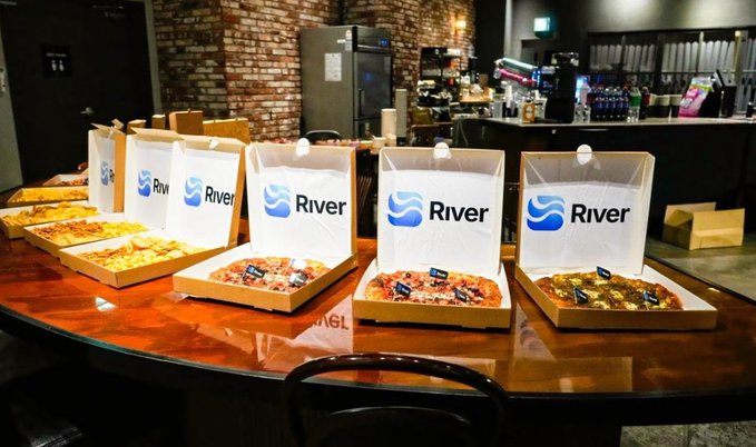 The image shows a table covered with several pizza boxes, each containing a different type of pizza. There are four pizzas visible, including one that has "RIVER" written on it. The table appears to be located inside a restaurant or a similar establishment where people can enjoy their meal and socialize.