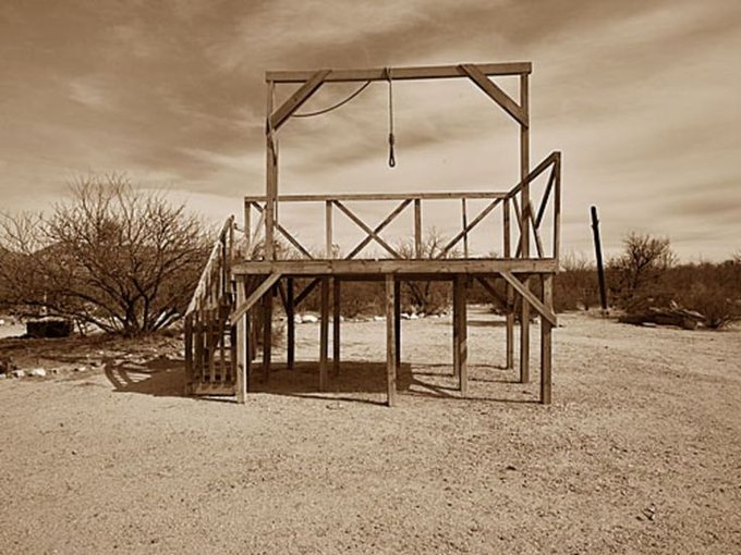 A sepia-toned photograph of a wooden gallows structure with a hanging noose, standing alone in a barren desert landscape with sparse brush.
