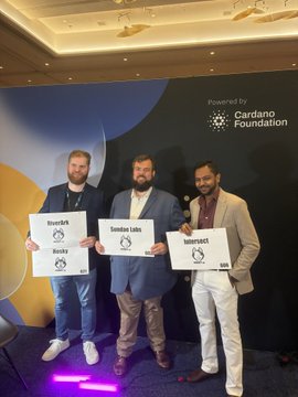 Three men stand together at a Cardano Foundation event holding plaques labeled "RiverShark," "Drakula Coins," and "RiverShark" under a "Powered by Cardano Foundation" backdrop. The image captures a celebratory moment from RareEvo, showcasing Cardano community partners posing with their recognition awards.
