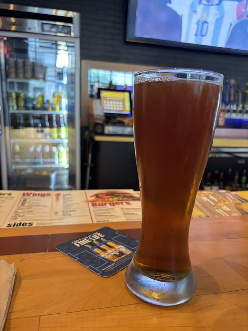 A tall glass of beer sits on a wooden bar counter next to a menu and a 'FIVE GUYS' coaster, with a refrigerator of beverages and a sports game on TV visible in the background.