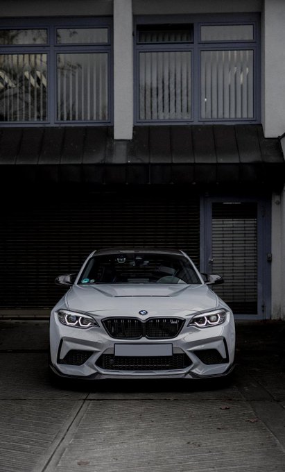 A head-on photograph of a silver BMW M2 parked in front of a building with a closed roller shutter and windows above.