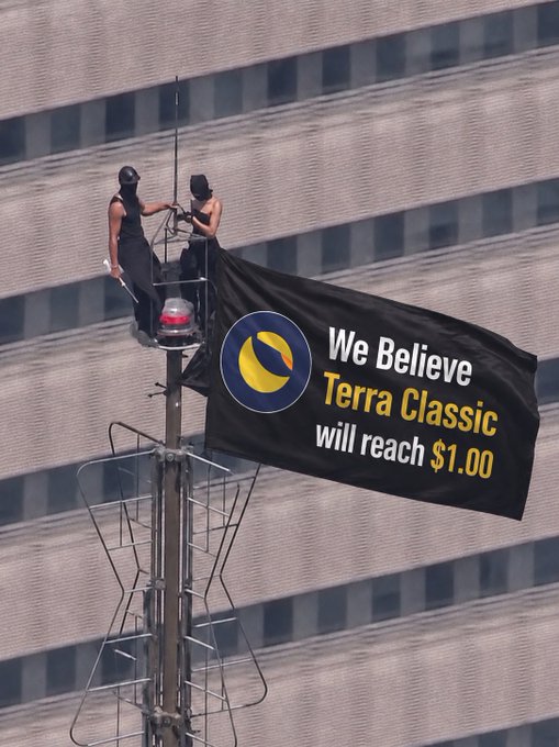 Two people stand atop a tall pole, unfurling a black banner that reads "We Believe Terra Classic will reach $1.00," featuring the $LUNC logo. The image visually echoes the post's rallying cry to push the cryptocurrency back to the $1 milestone.