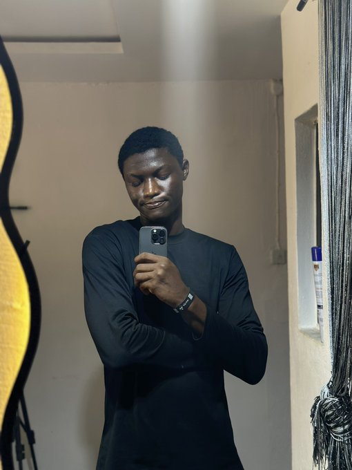 A young man in a black long-sleeved shirt smiles while taking a mirror selfie with his smartphone in what appears to be a room with a guitar visible on the left.