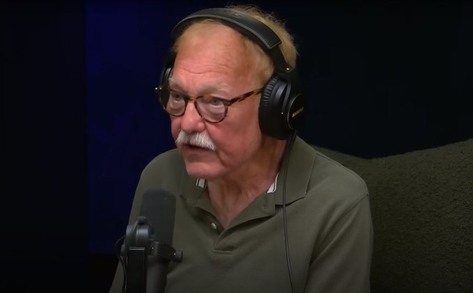 An elderly man with a white mustache, glasses, and headphones sits in front of a microphone, appearing to be recording a podcast or interview.