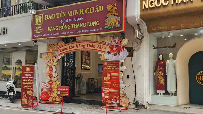 The image shows a store front with red banners, including one that says "BÐT TÜ MINH CHAU" and another that reads "VAN GØCH THA". The store appears to be located on a street in Vietnam, as indicated by the presence of a motorcycle parked outside. A person is also visible near the store front, possibly browsing or waiting for someone else.
