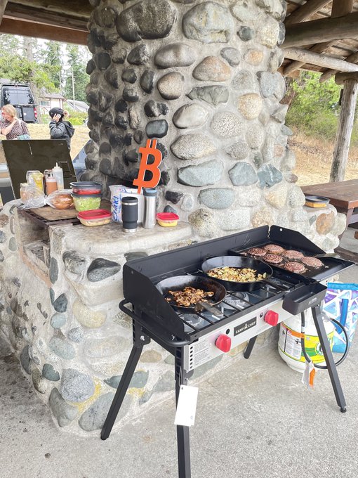 An outdoor scene showing a Blackstone griddle cooking food on a stone patio, with an orange Bitcoin 'B' decoration mounted on a stone pillar.