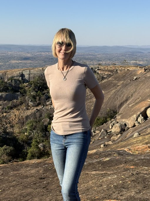 A woman with blonde hair and glasses posing on a rocky outcrop with a scenic valley landscape stretching into the distance behind her.